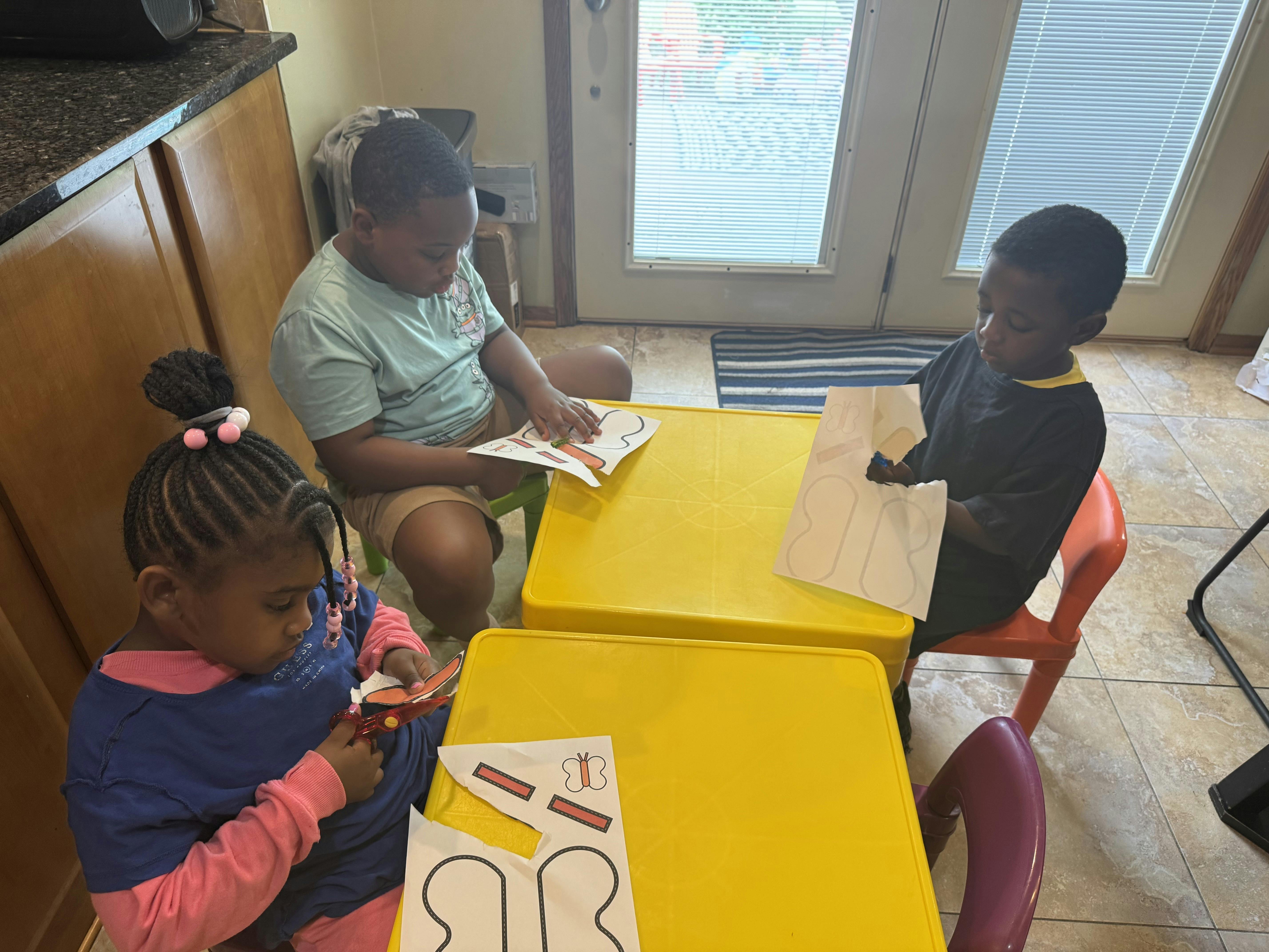 Three children working on paper craft projects at small yellow tables