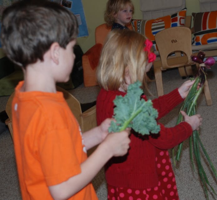 Child in orange shirt holding kale, child in red shirt holding green vegetables