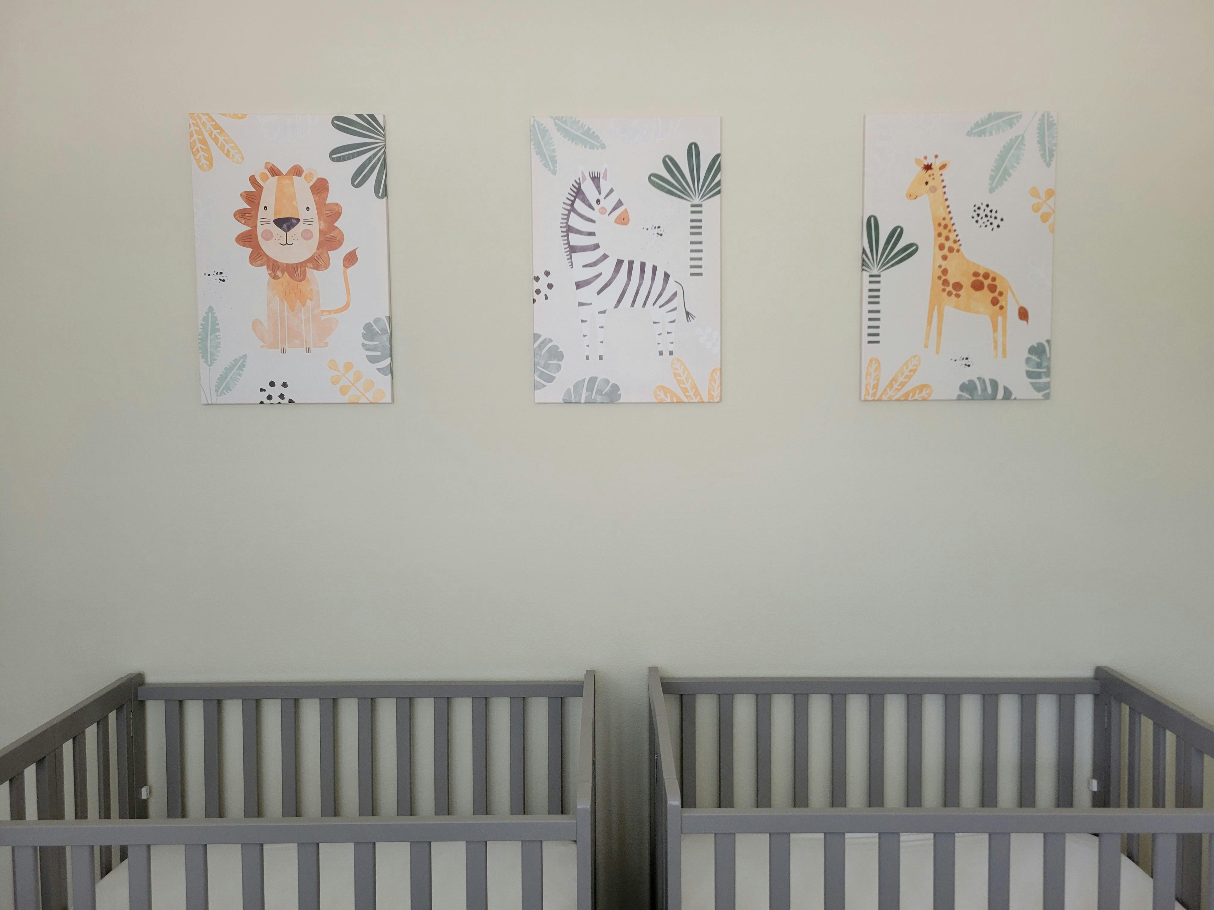 Two cribs below three animal-themed pictures on a wall