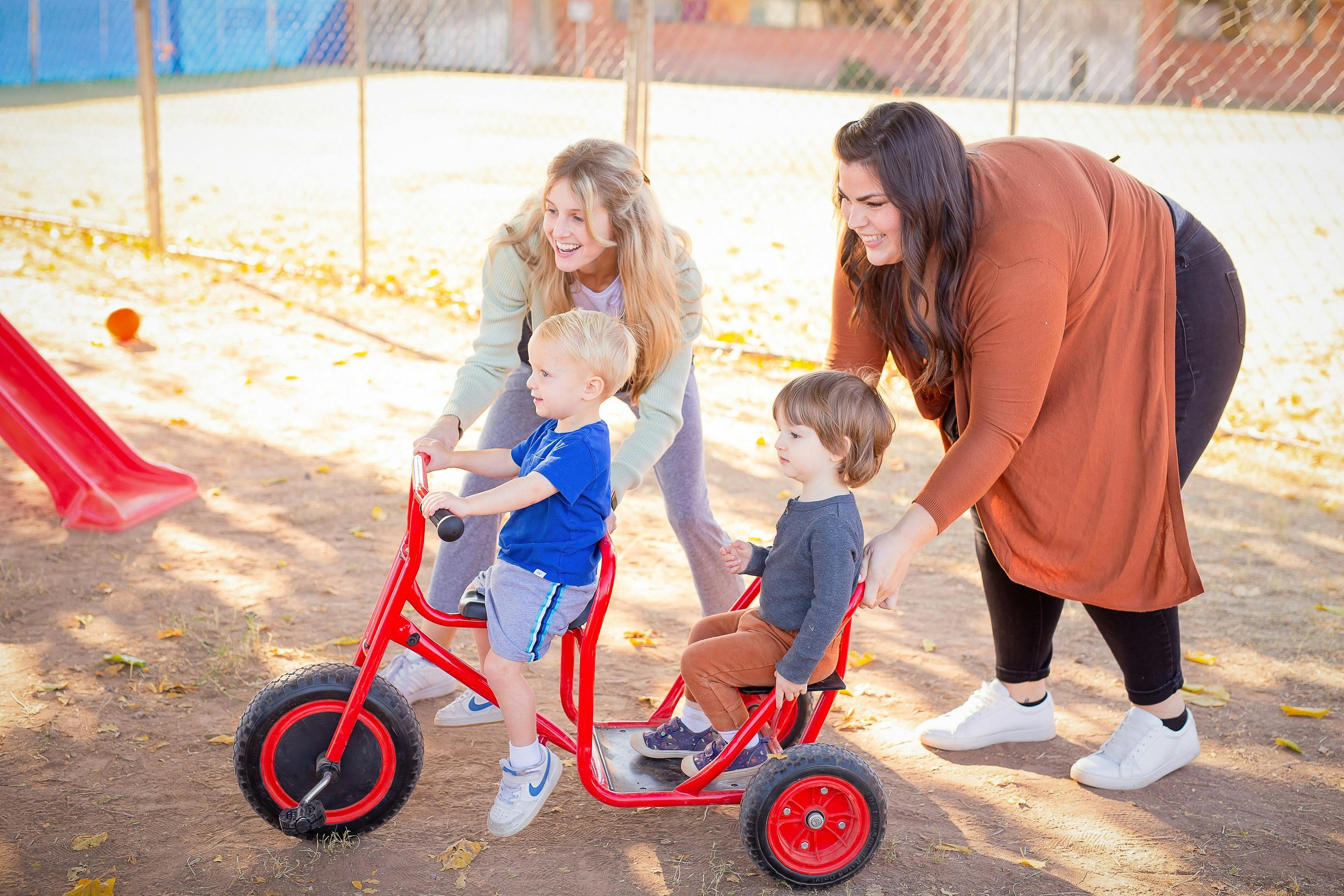Two people assisting two children riding a red tandem tricycle on a playground
