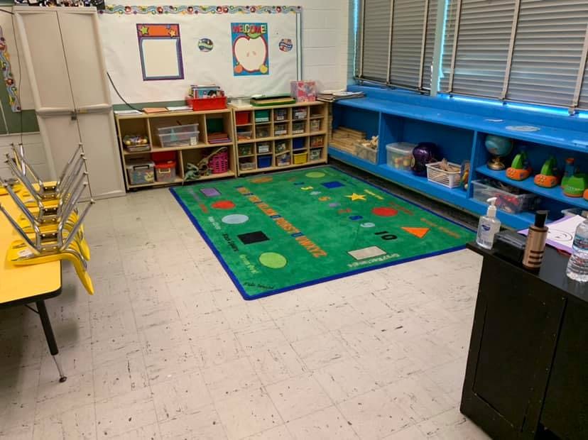 Classroom with a green rug, shelves, and a yellow table with stacked chairs