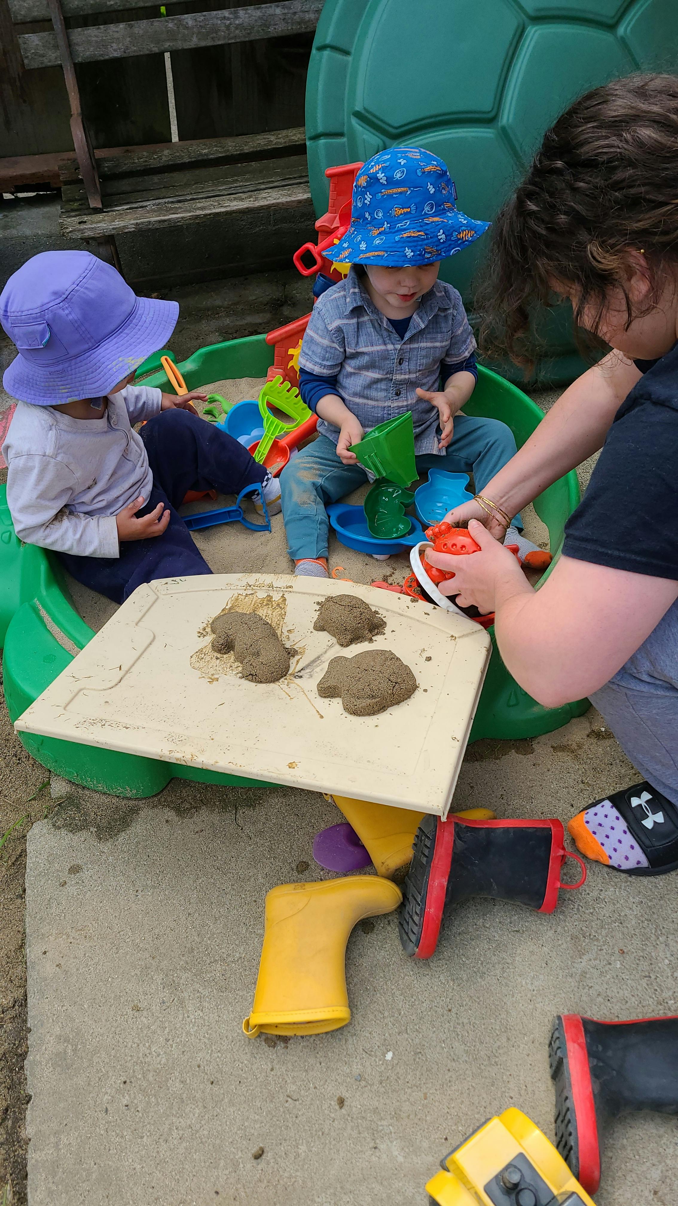 Two children and an adult playing in a sandbox with sand toys