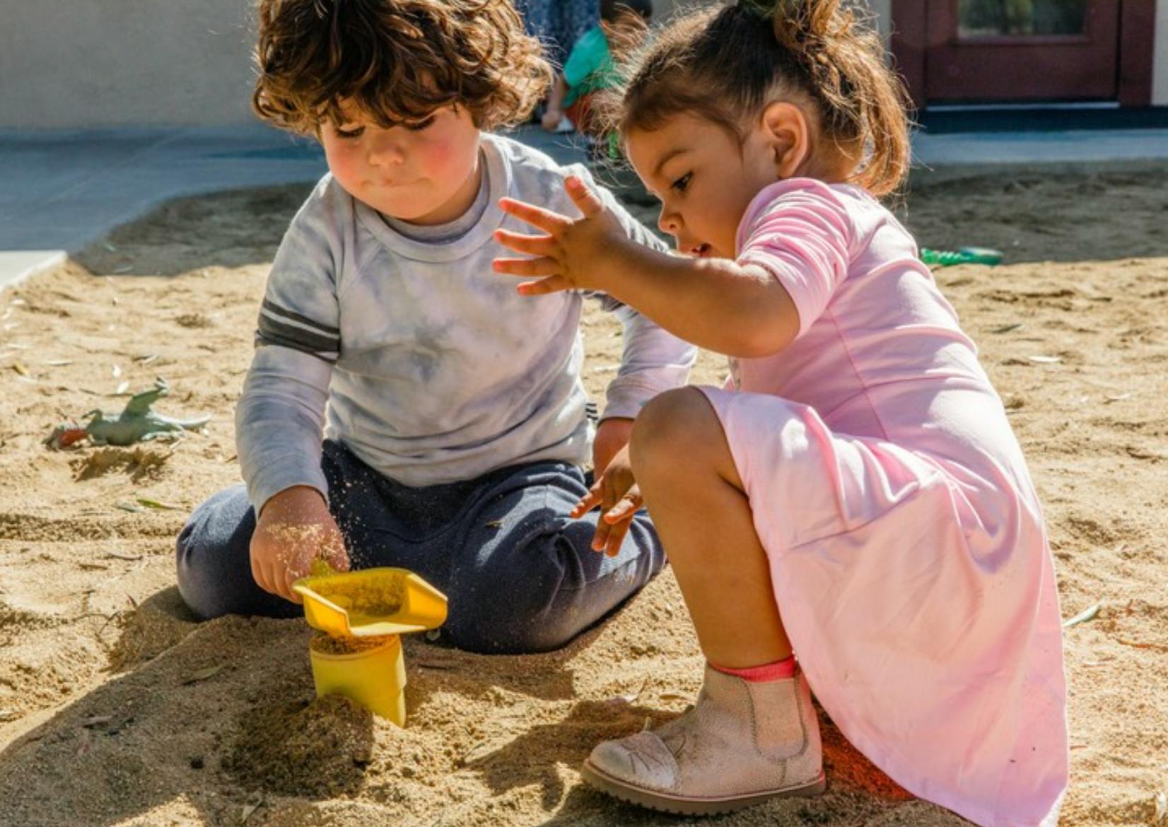 Two children playing in a sandbox with a yellow scoop