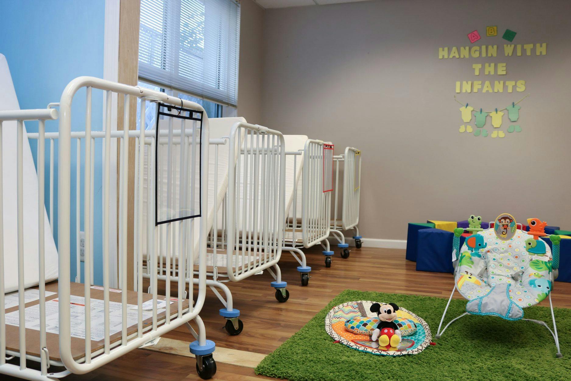 Row of white cribs in a room with a play mat and infant bouncer