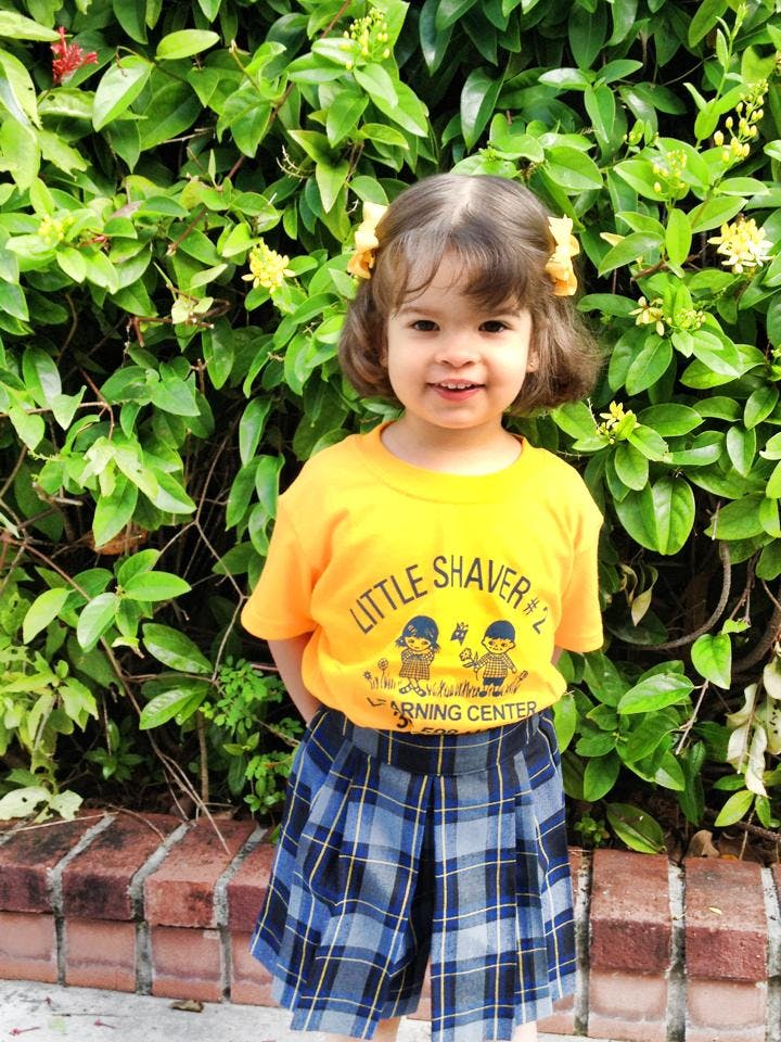 Child in yellow shirt and plaid skirt standing in front of green foliage