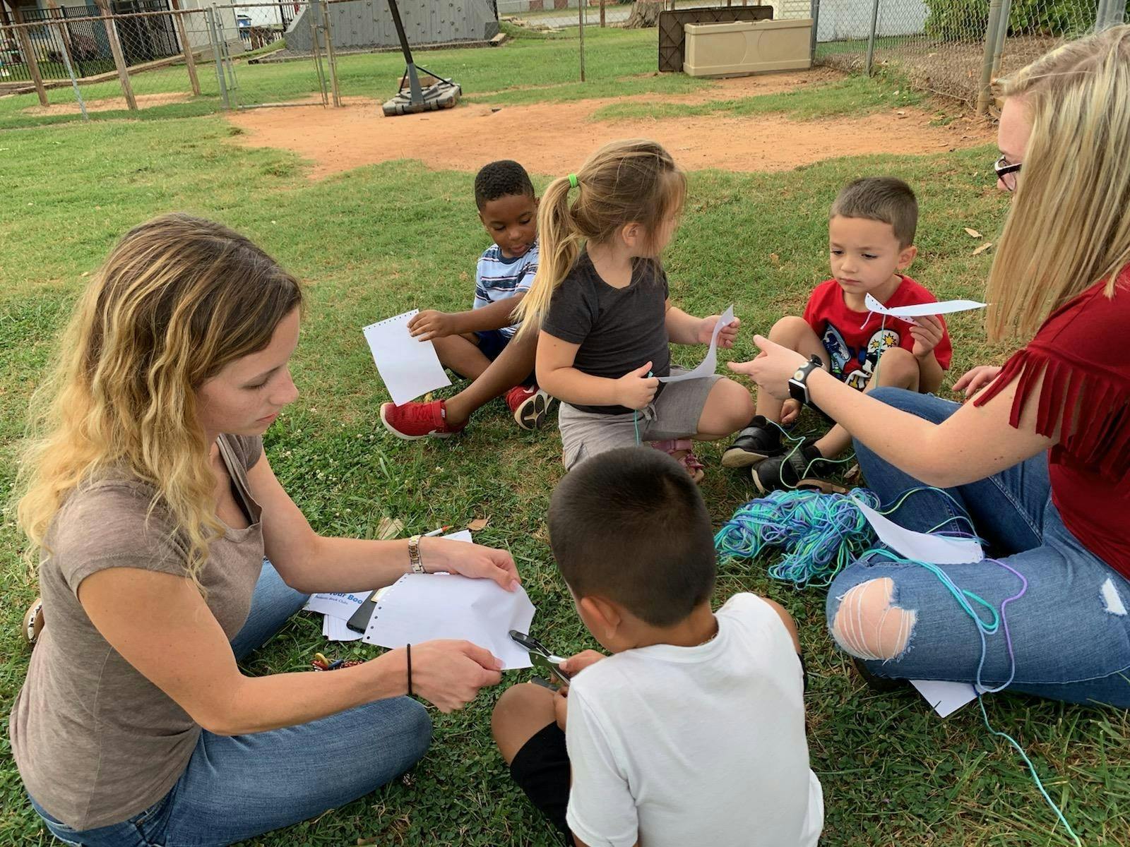 Two adults and four children sitting on grass, working with paper