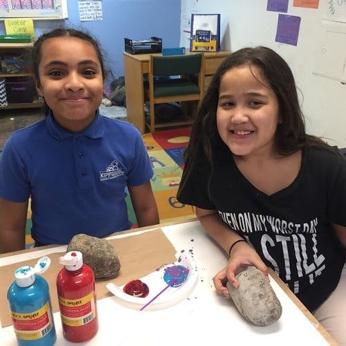 Two children sitting at a table with paint and rocks