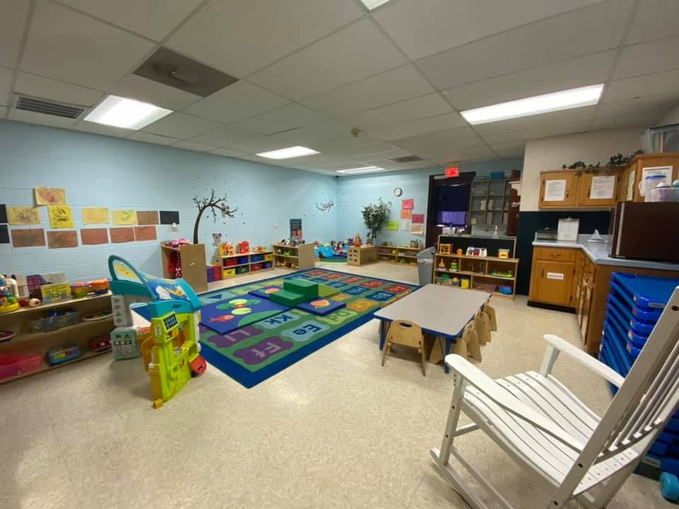 An empty classroom with a colorful play rug, small tables, and shelves of toys