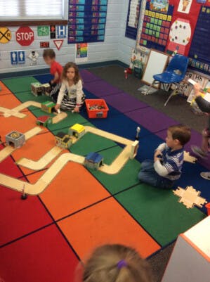 Children playing with toy cars and tracks on a colorful rug in a classroom