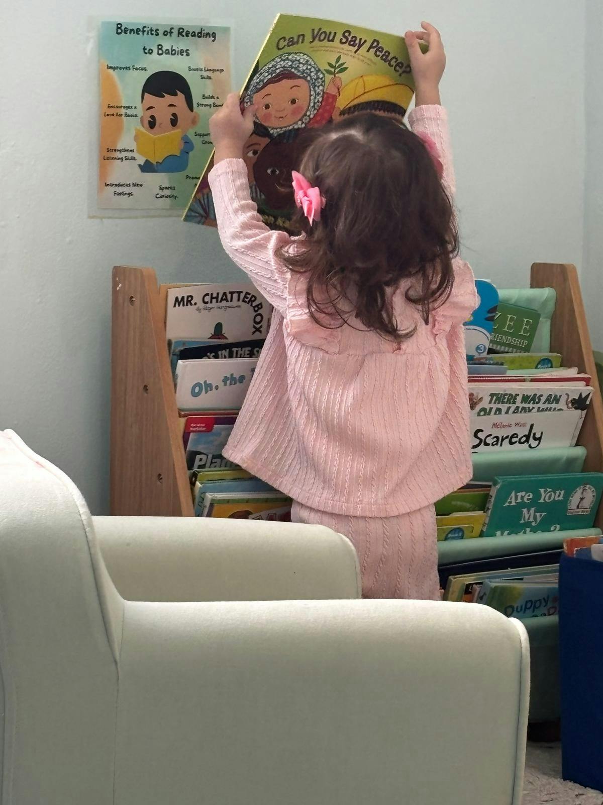 Child standing, holding a book titled "Can You Say Peace?" near a bookshelf