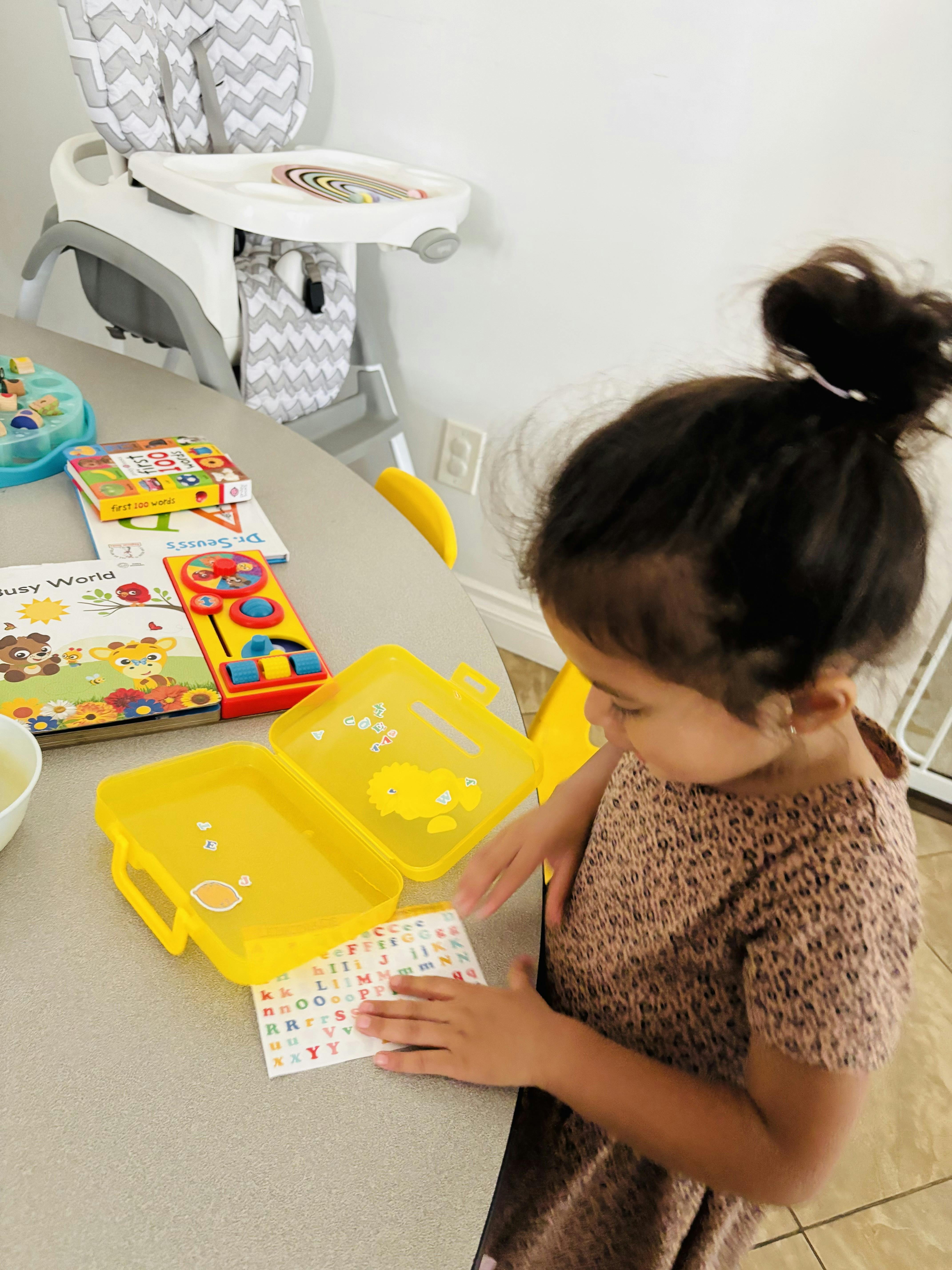 Child placing alphabet stickers on a yellow case next to several books and toys