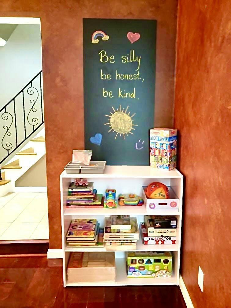 Chalkboard with "Be silly, be honest, be kind" above a white bookshelf with books and toys