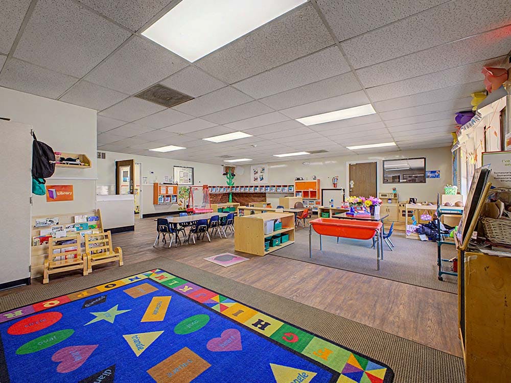 An empty classroom with a colorful rug on the floor and small tables and chairs