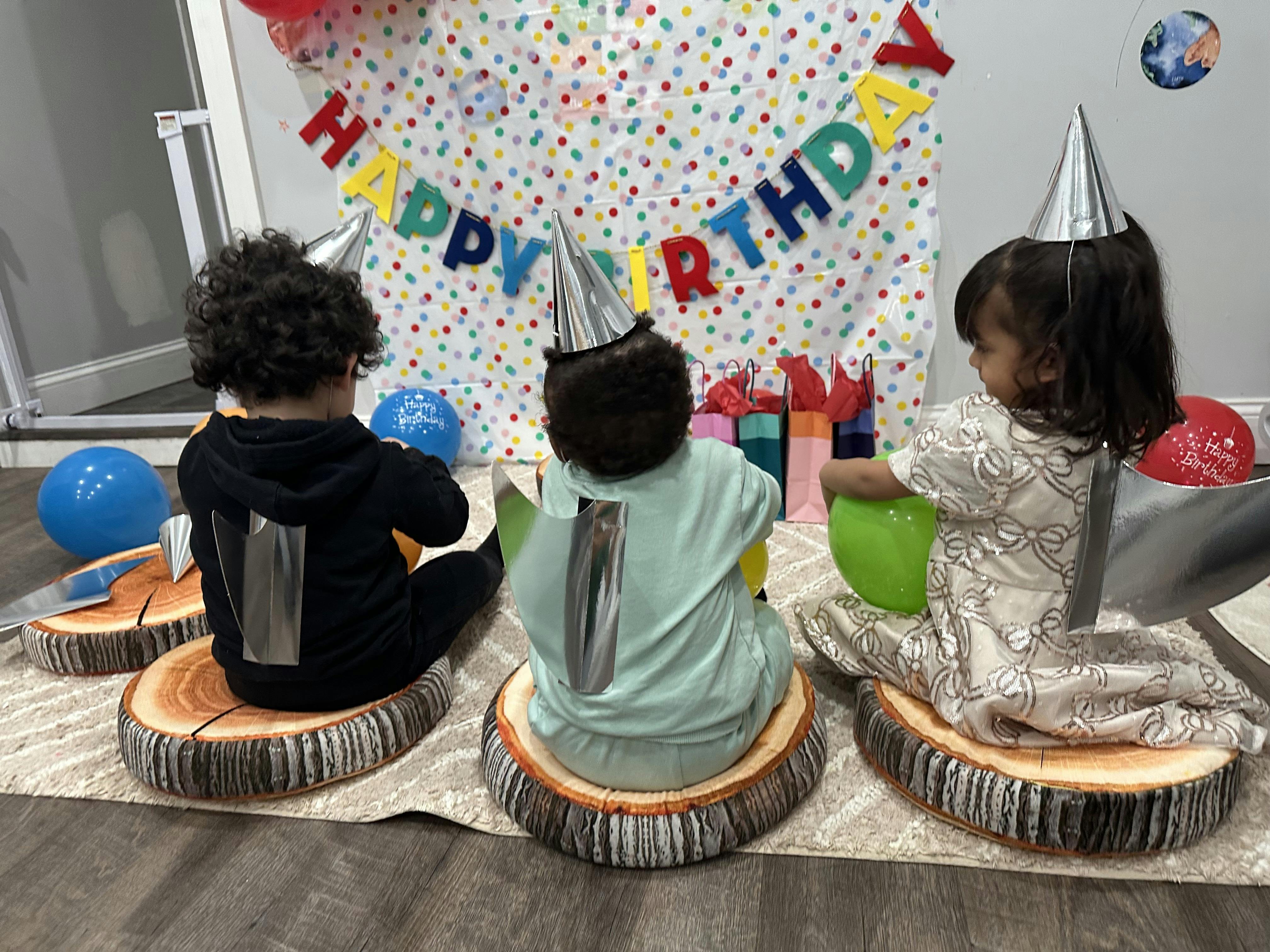 Three children in party hats sitting on circular tree stump cushions, facing a "Happy Birthday" banner