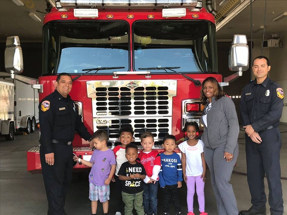 Two people in uniform and one person with several children in front of a fire truck