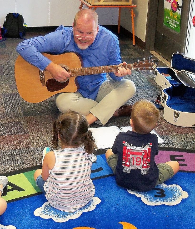 Person playing guitar for two children in a classroom