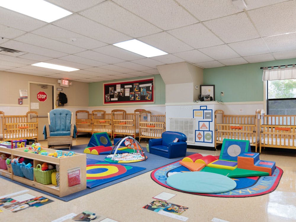 A colorful daycare room with several cribs, soft play equipment, and shelves of children's toys