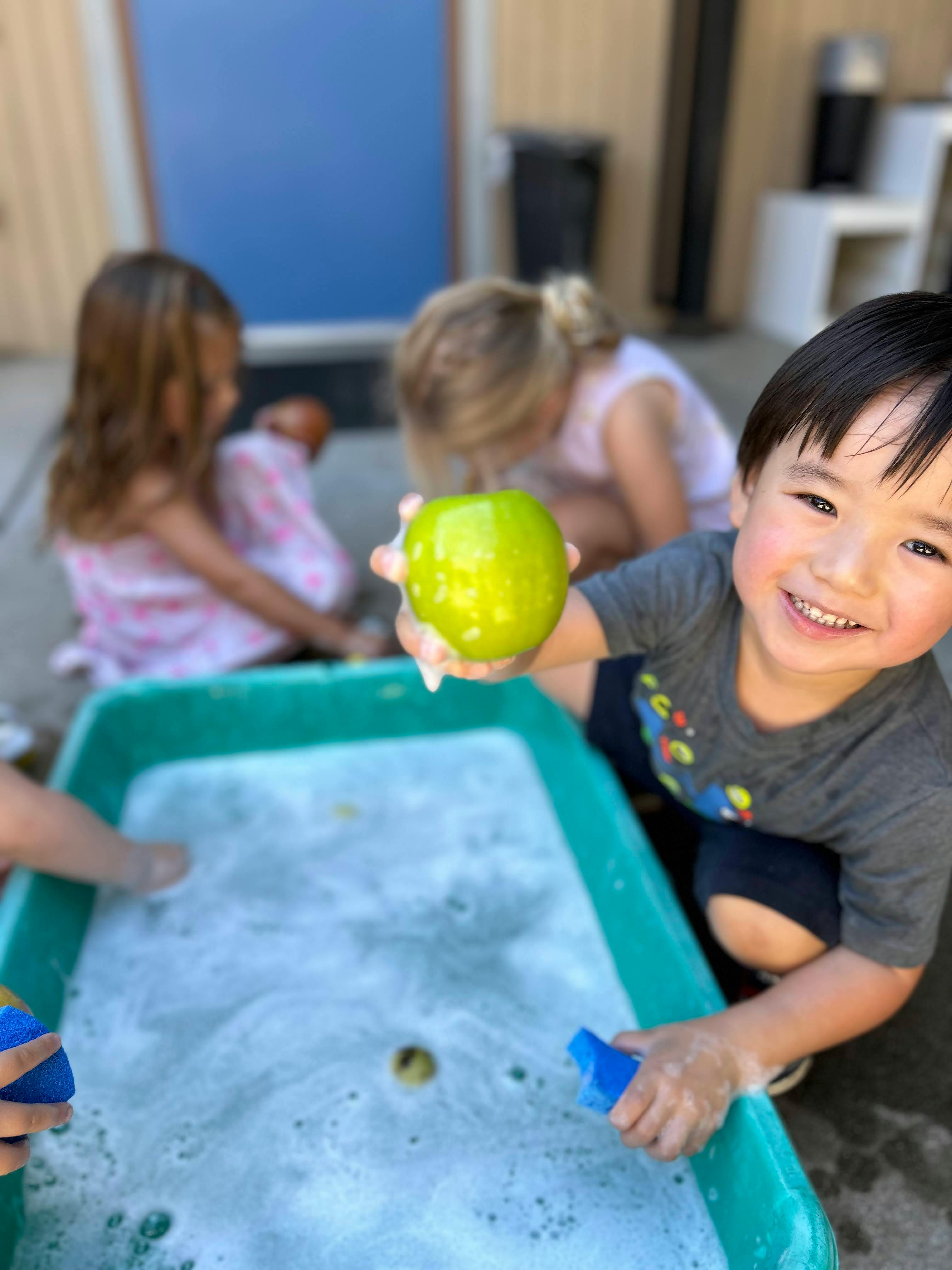 Child holding a green apple over a tub of soapy water