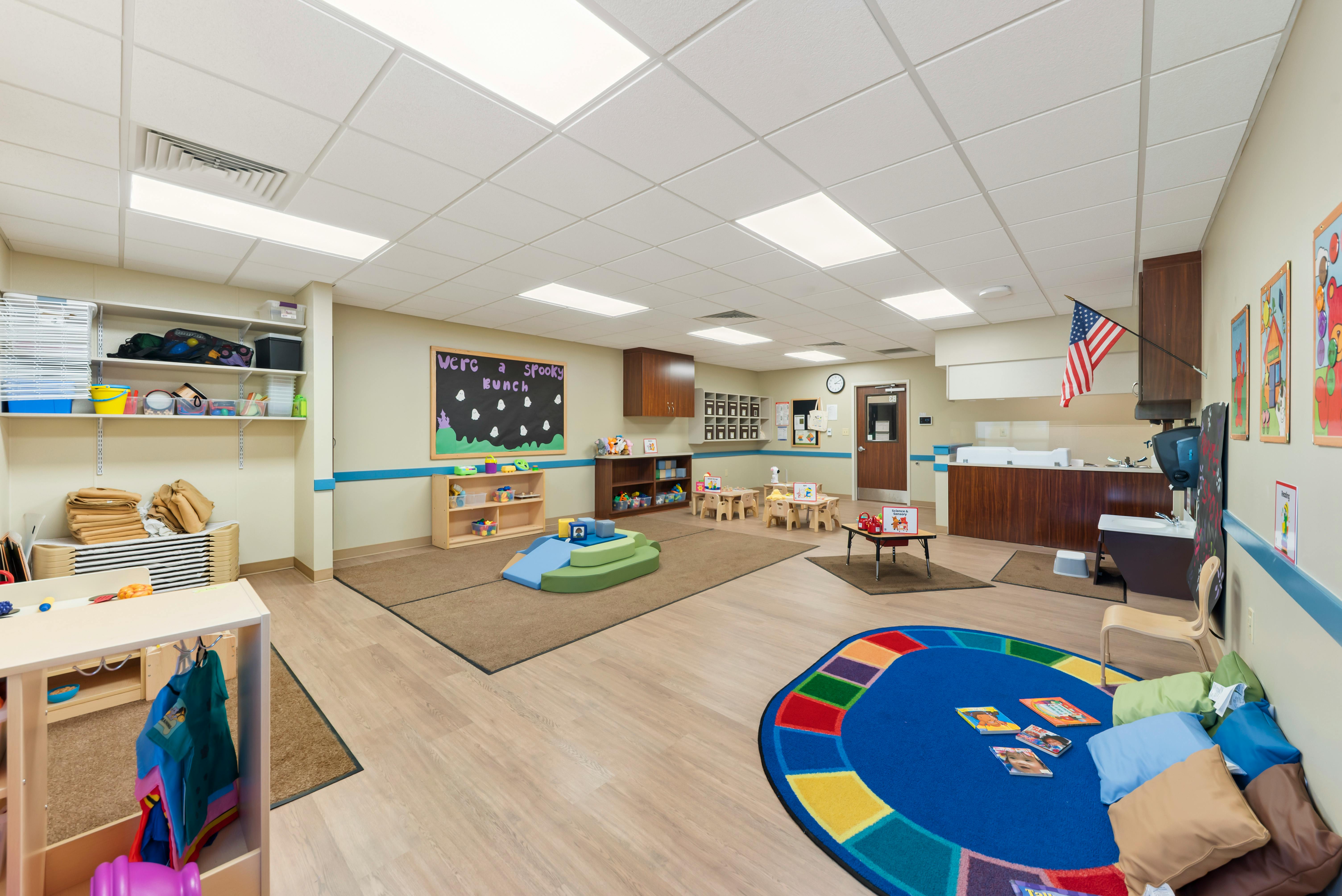 Classroom with small tables and chairs, shelves with toys, and a colorful rug