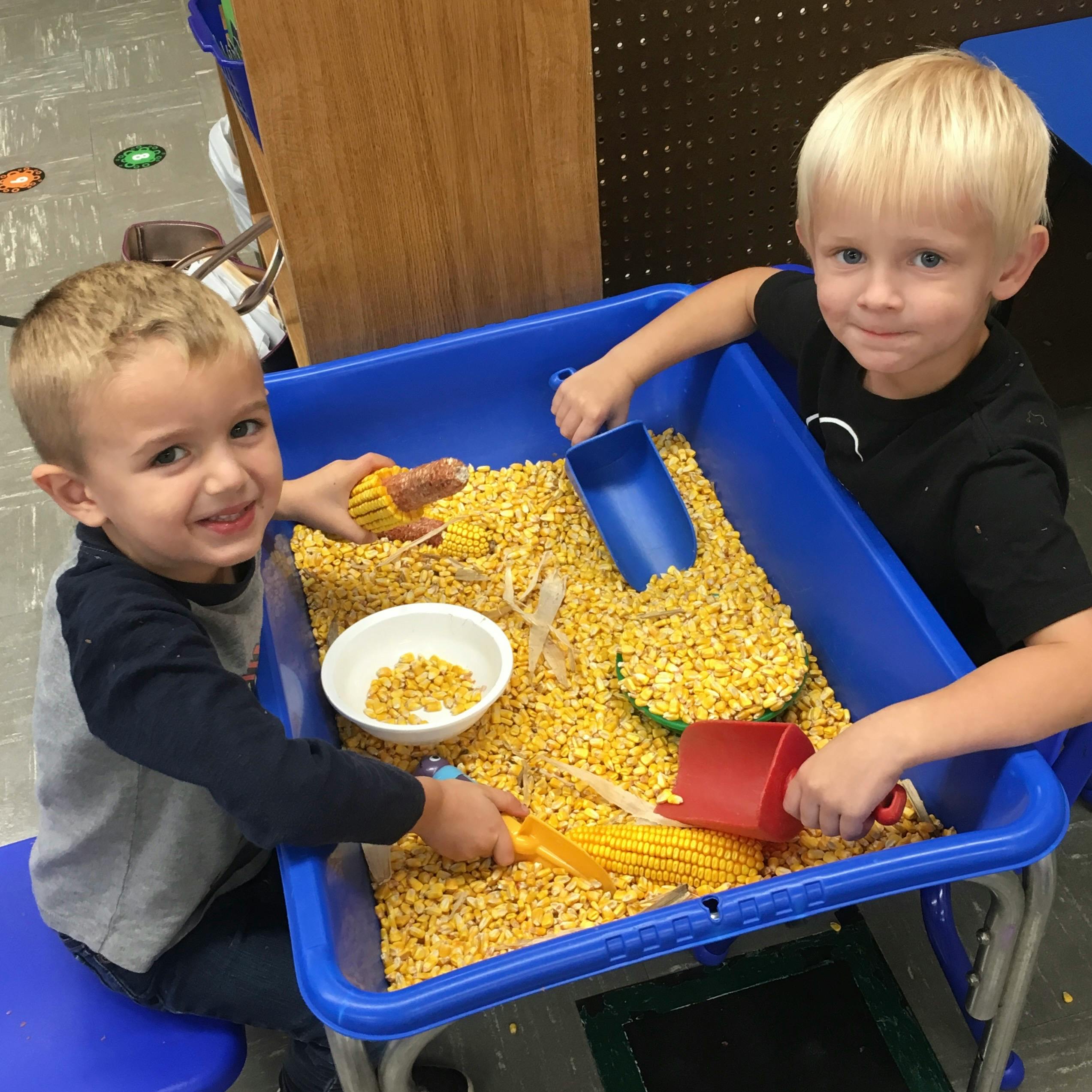 Two children playing with corn kernels in a blue sensory bin