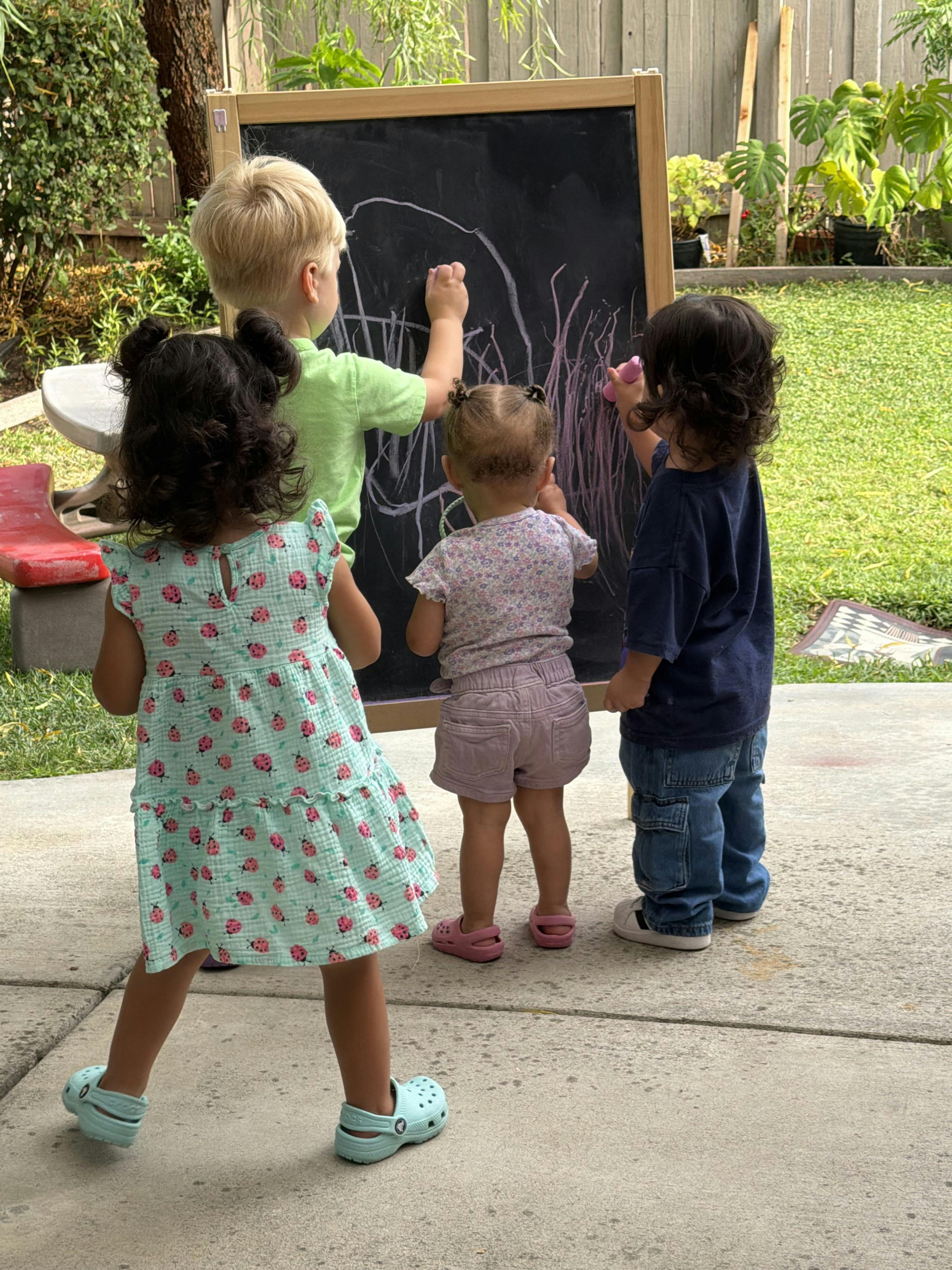 Four children drawing on a chalkboard in a backyard