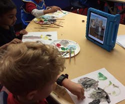 Children painting at a table with a tablet displaying an image