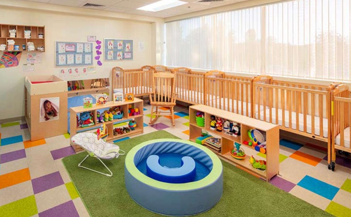 Nursery room with multiple cribs, shelves of toys, and a soft play area