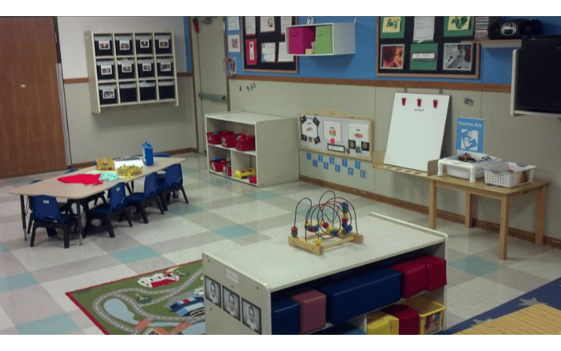 empty preschool classroom with tables, chairs, shelving, and play rug