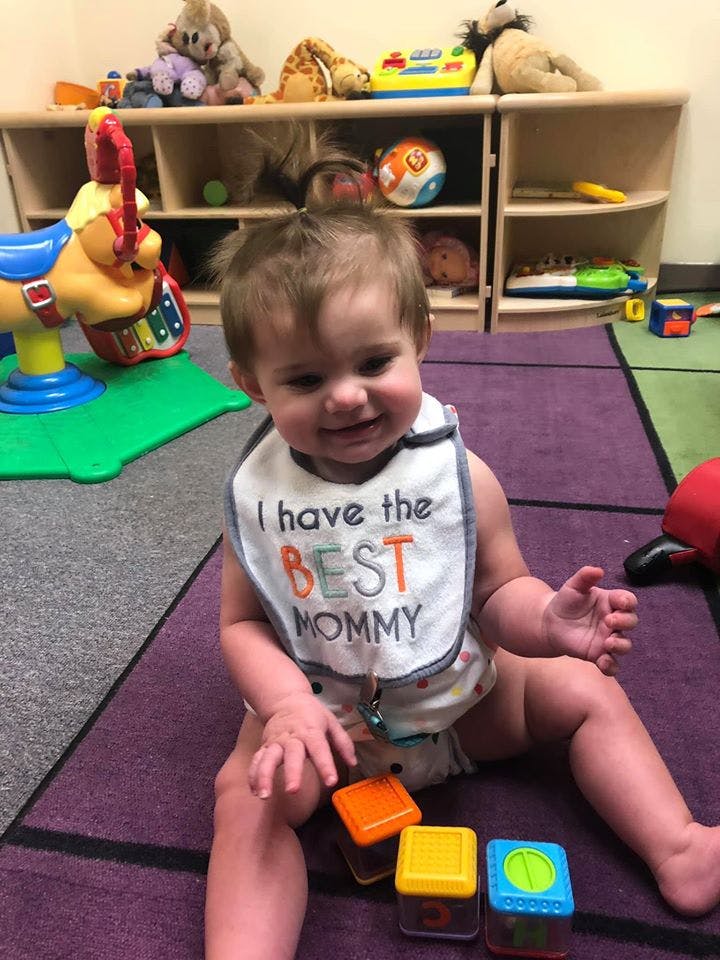 Child wearing a bib that says "I have the BEST MOMMY" sitting on a rug with toys