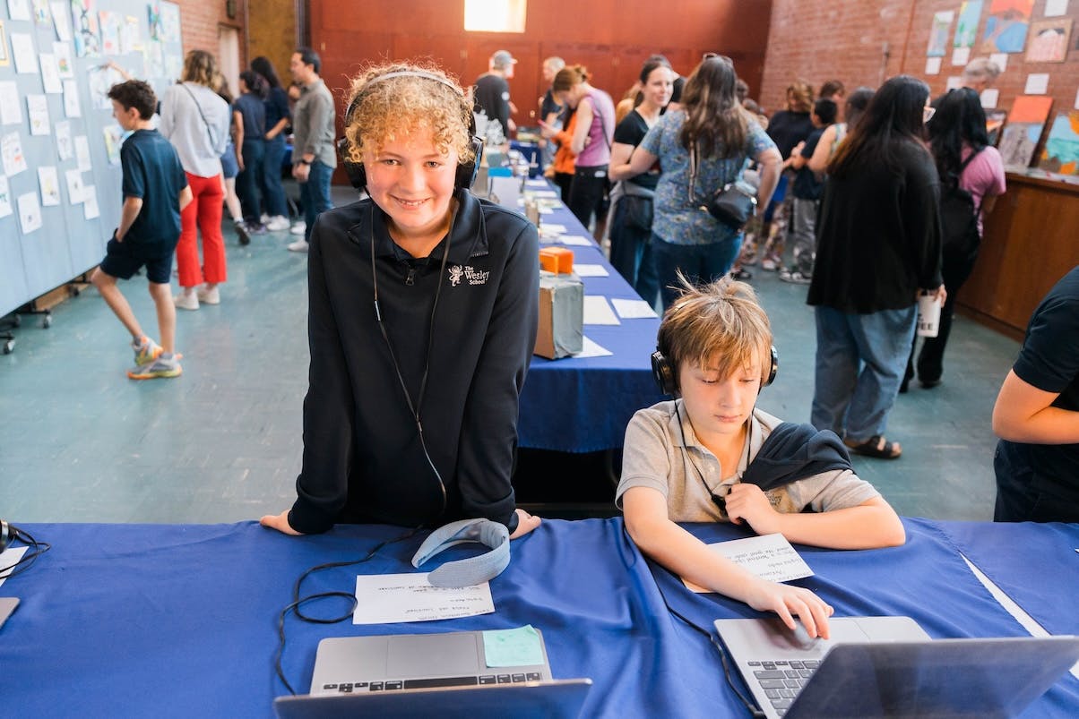 Two children with headphones at a table with laptops, one smiling at the camera