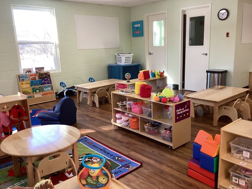 A brightly lit classroom with small wooden tables, chairs, and shelves of toys