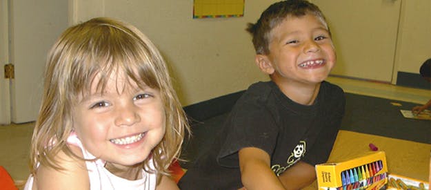 Two children smiling, one with a box of markers