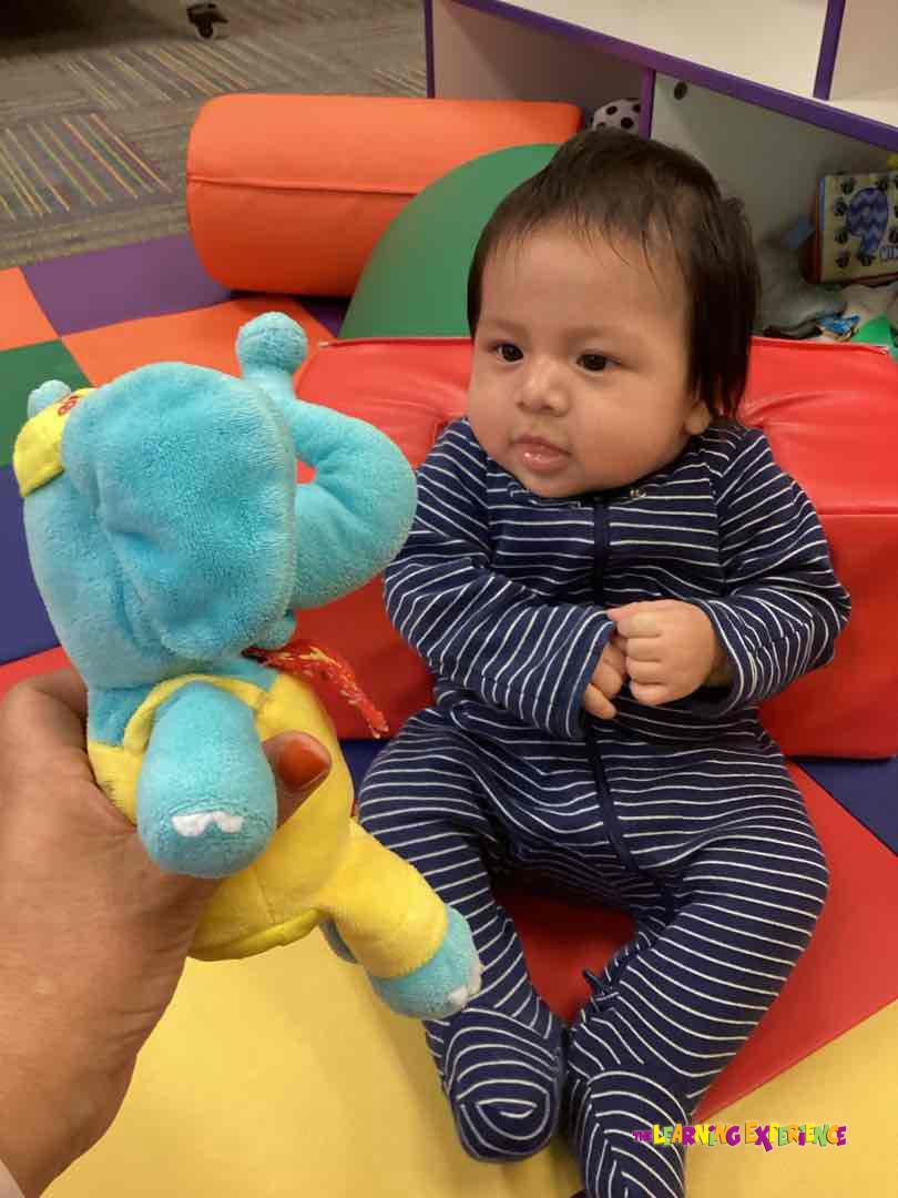 Person holding a blue stuffed animal next to a baby sitting on a play mat