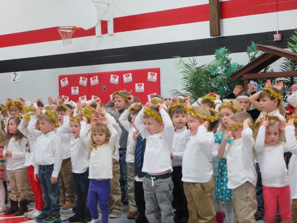 Group of children wearing crowns and white shirts, some with hands raised