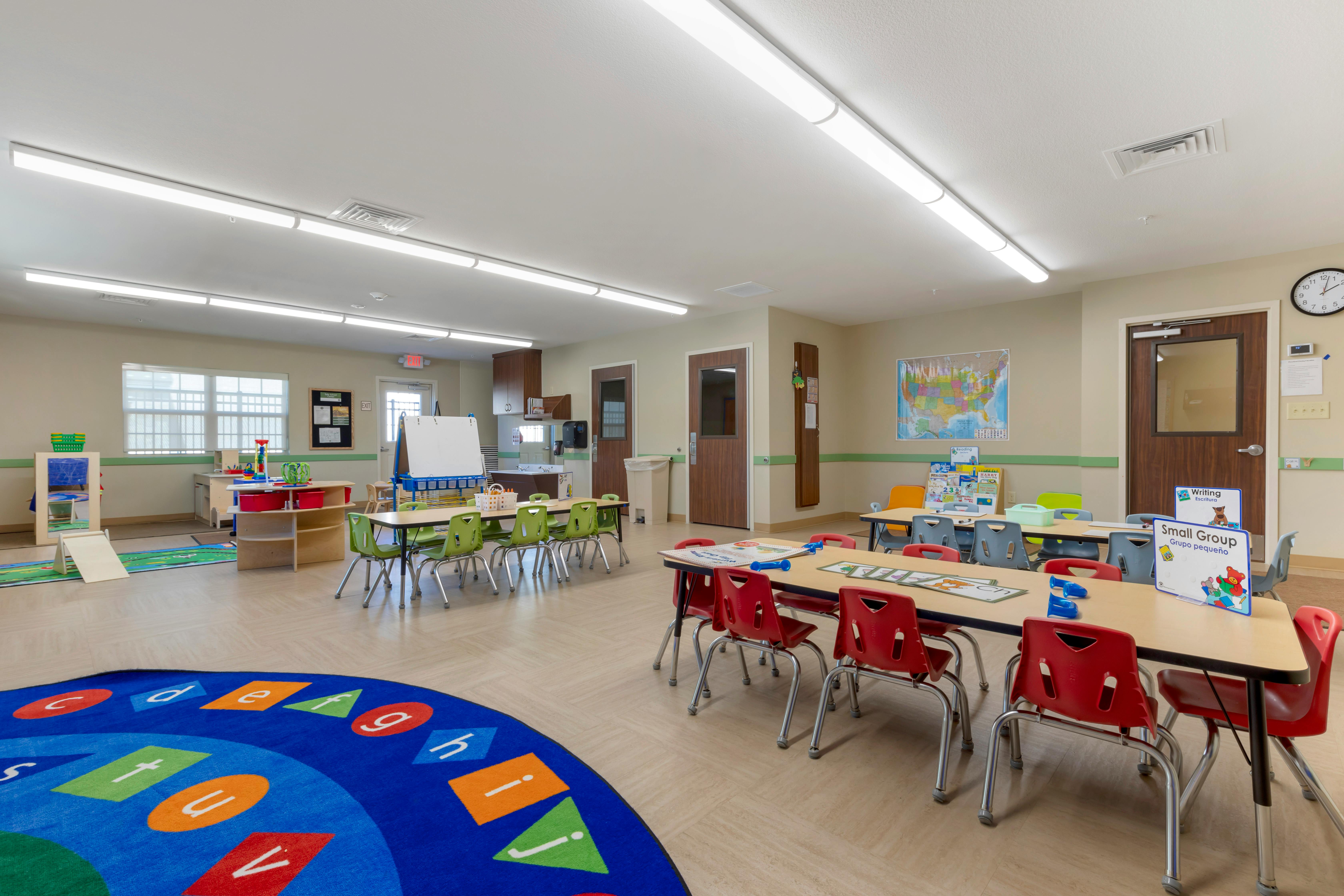 Classroom with small tables and chairs, a play area, and an alphabet rug