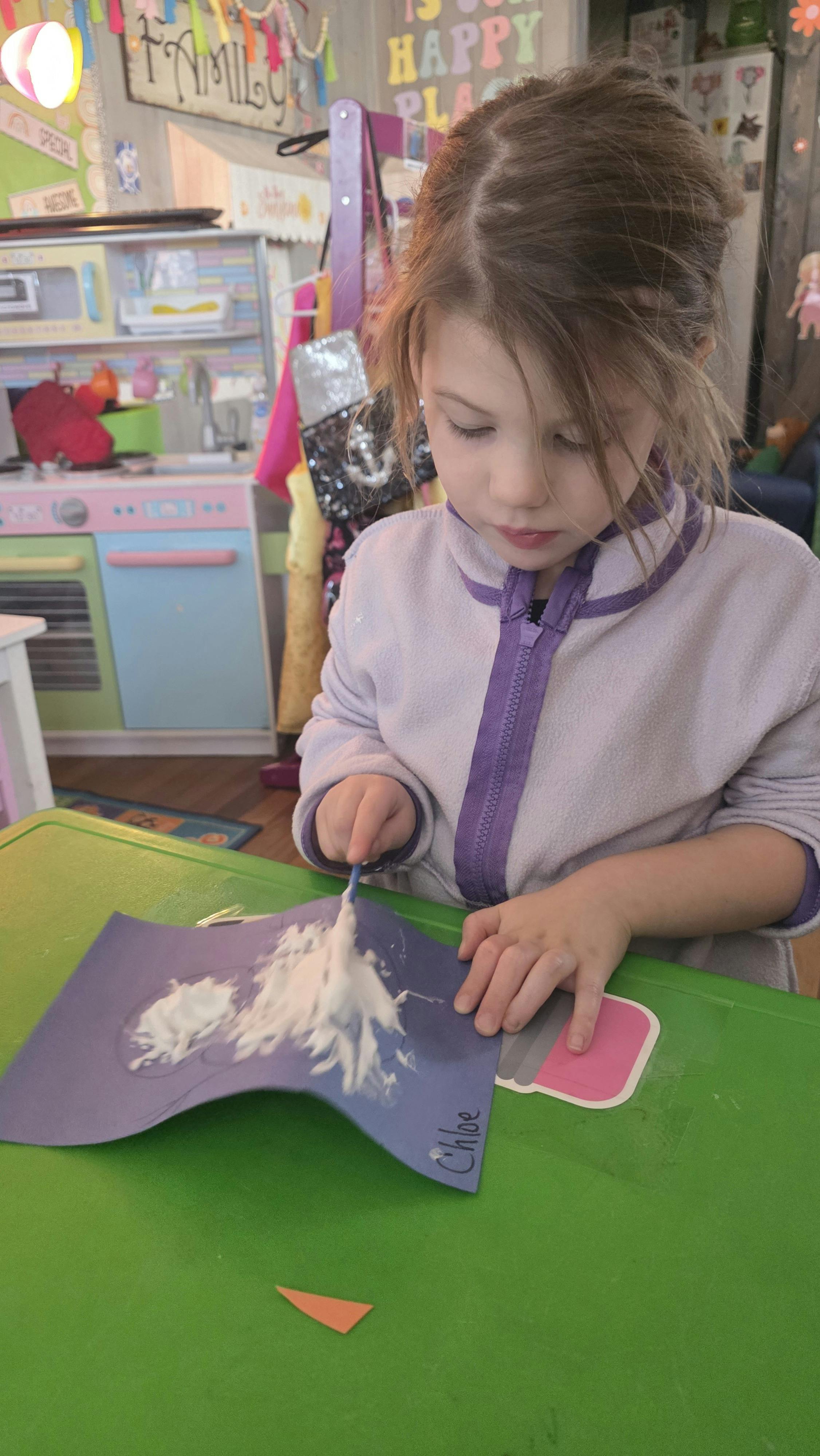 Child painting white glue on a blue paper cutout on a green table