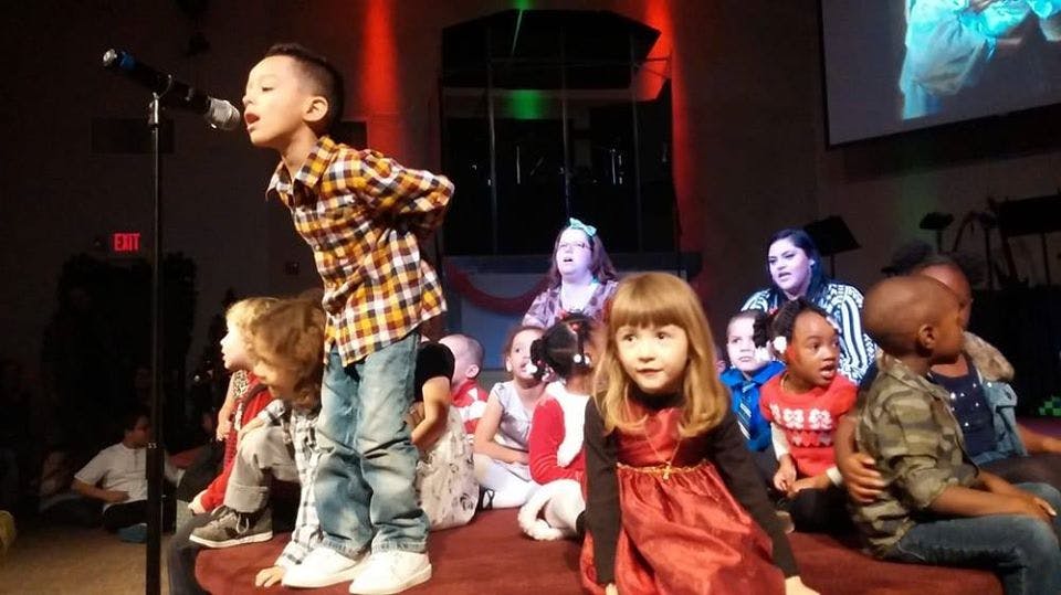 Child singing into a microphone on a stage with other children watching