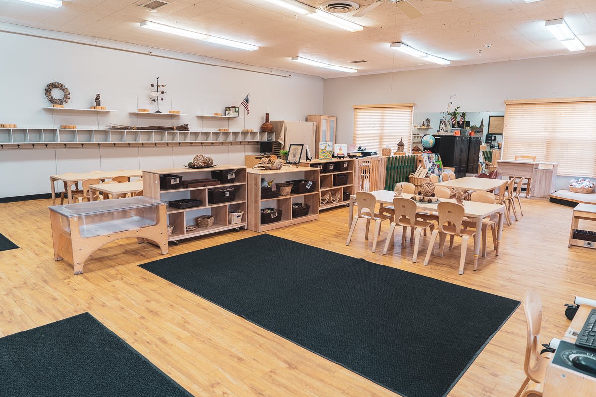 Classroom with small tables and chairs, shelves, and black rugs on a wood floor