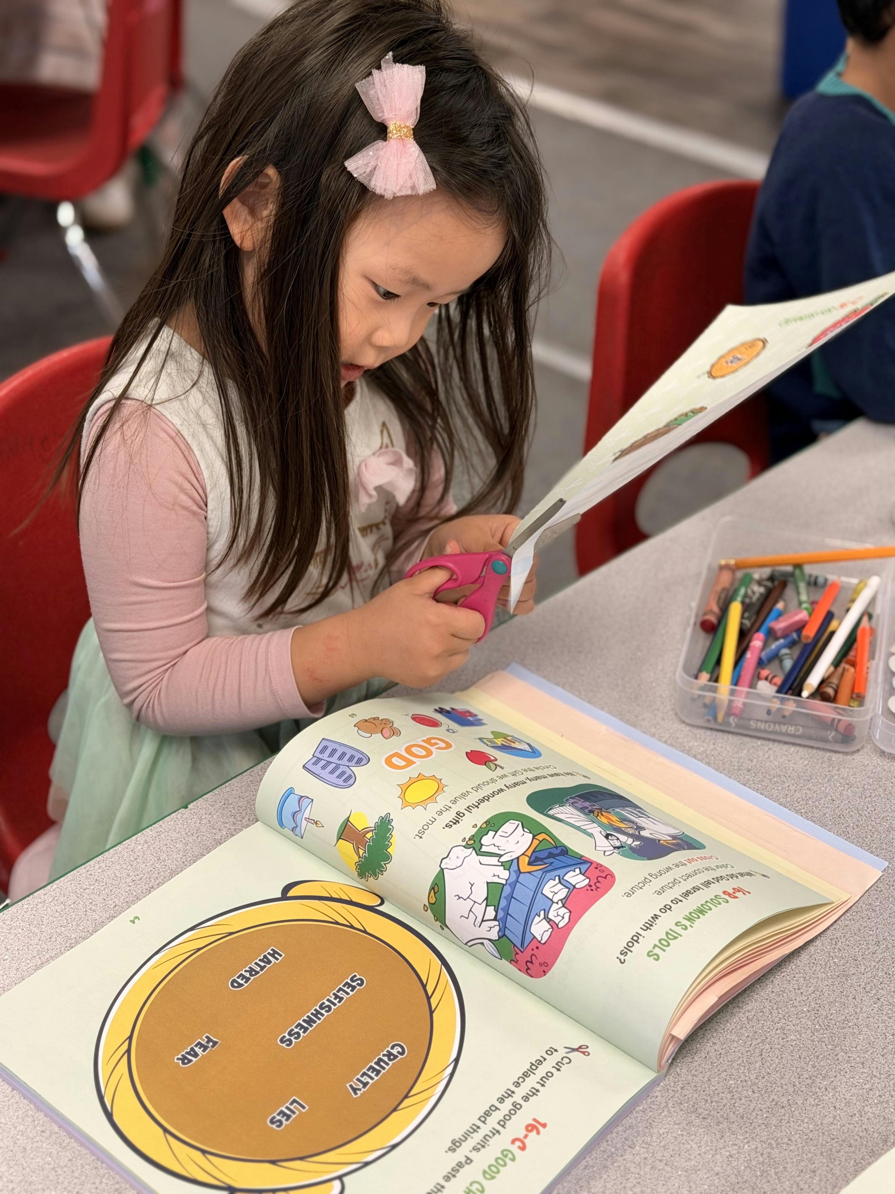 Child cutting paper with pink scissors while looking at a workbook