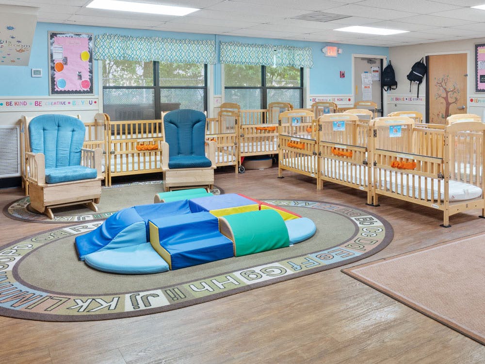 A brightly lit nursery room with multiple cribs, two rocking chairs, and soft play equipment