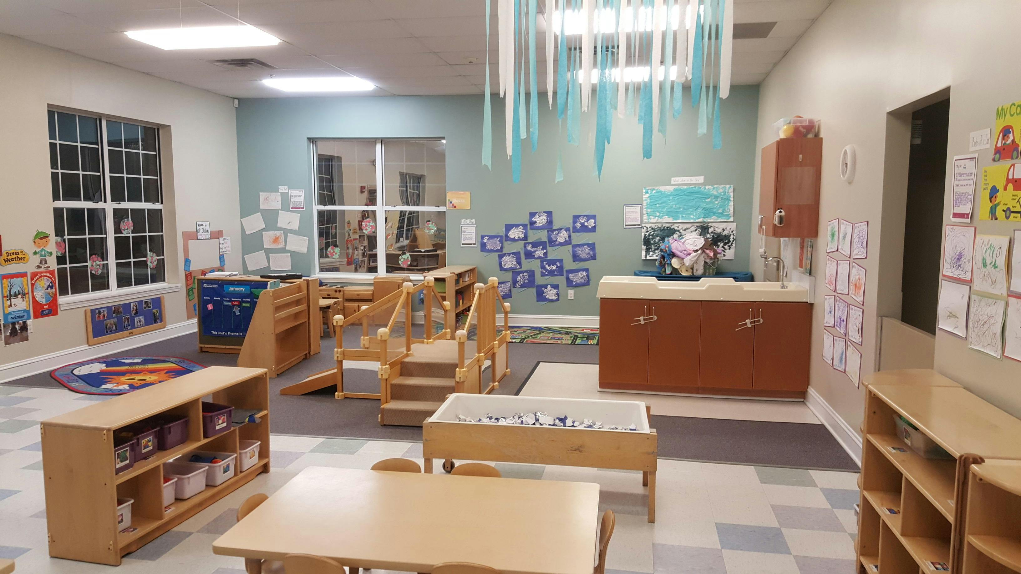 Empty classroom with small tables, chairs, shelving units, play structure, and artwork