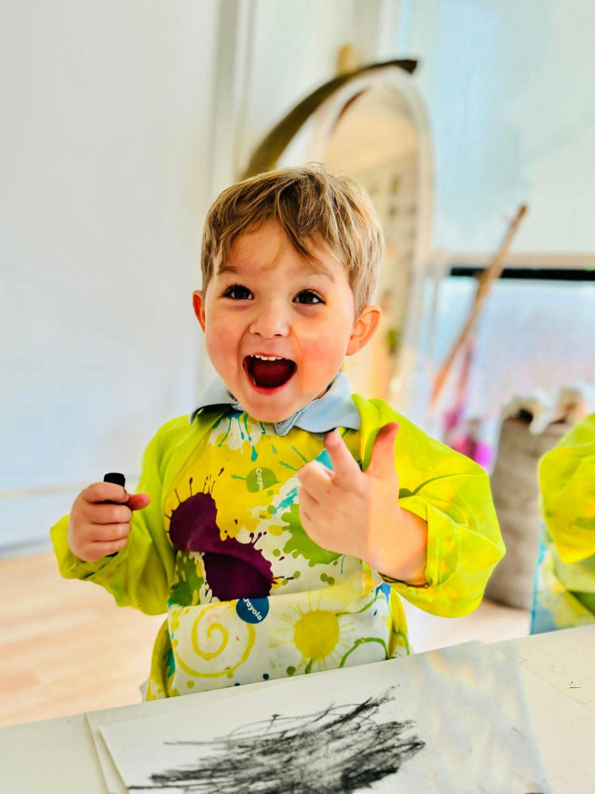 Child in a paint-splattered smock holding a marker and giving a thumbs up