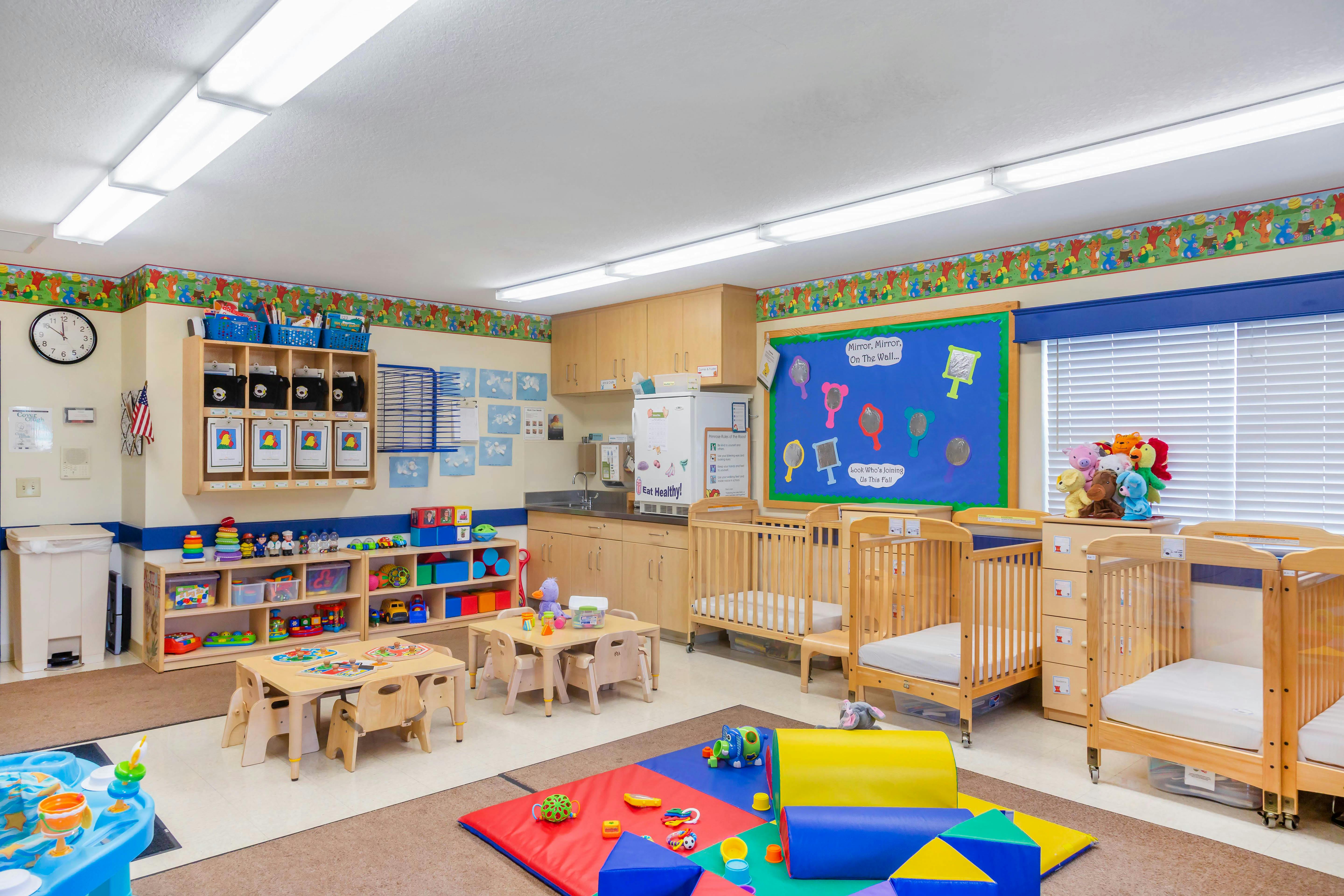 A classroom with cribs, small tables and chairs, shelves with toys, and a play mat