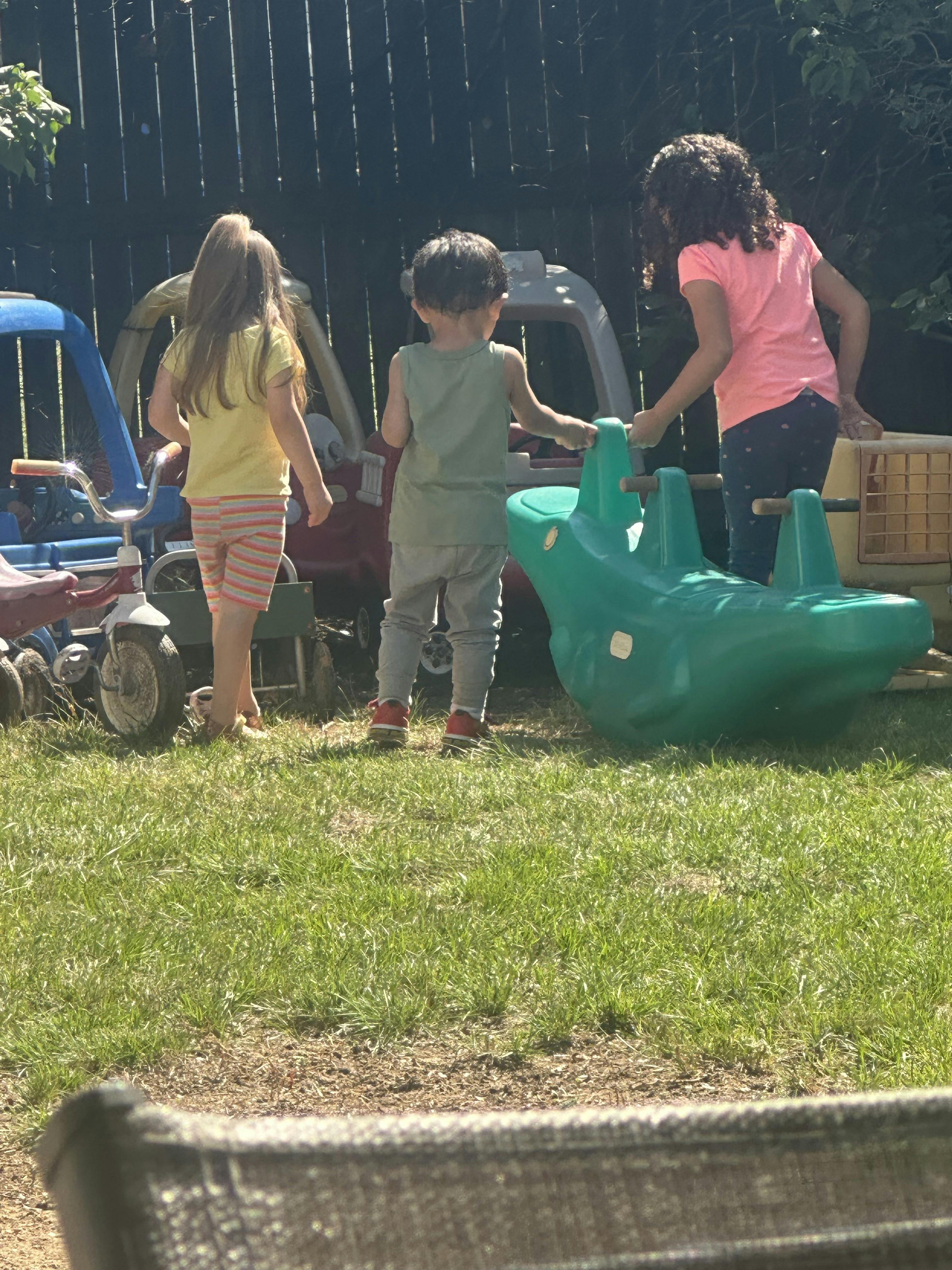 Three children playing with outdoor toys on grass, seen from behind
