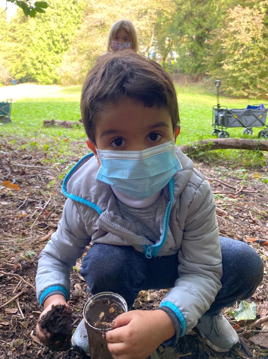 Child wearing a face mask, holding dirt and a container of mud