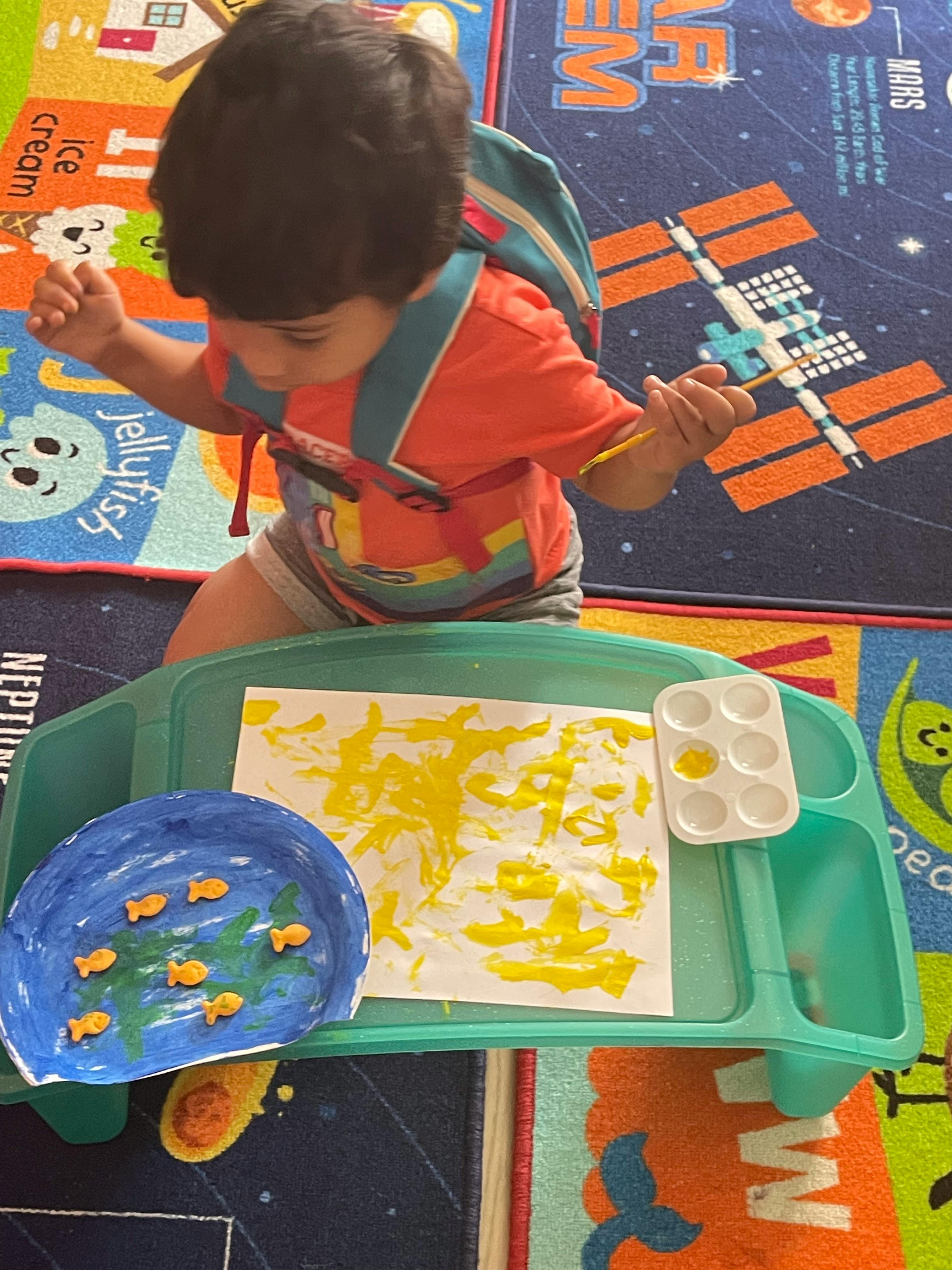 Child painting with yellow paint on a paper on a green tray
