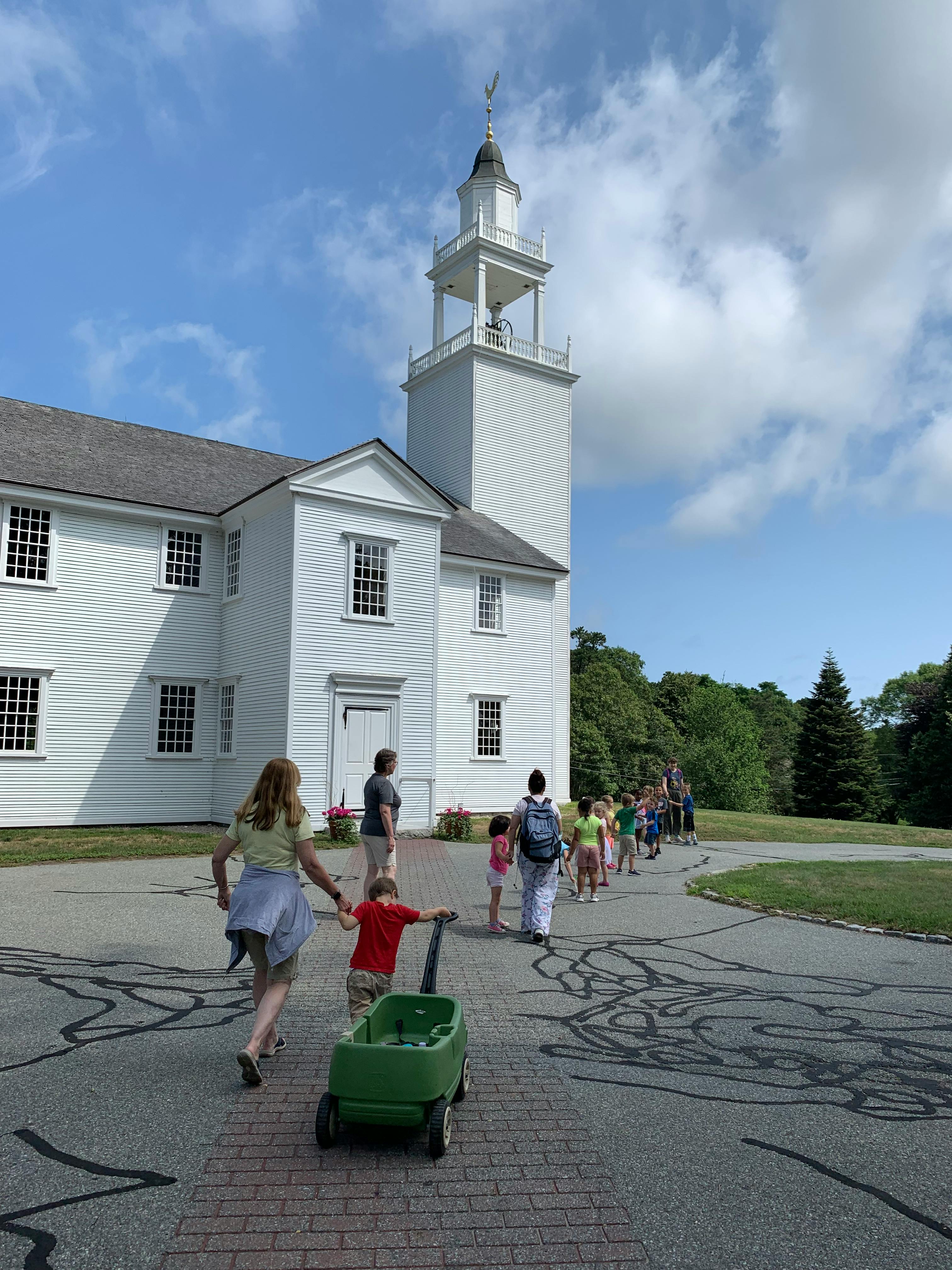 People and children walking on a path next to a white church with a steeple