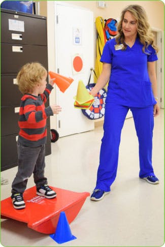 Person in scrubs with child on balance board holding cones
