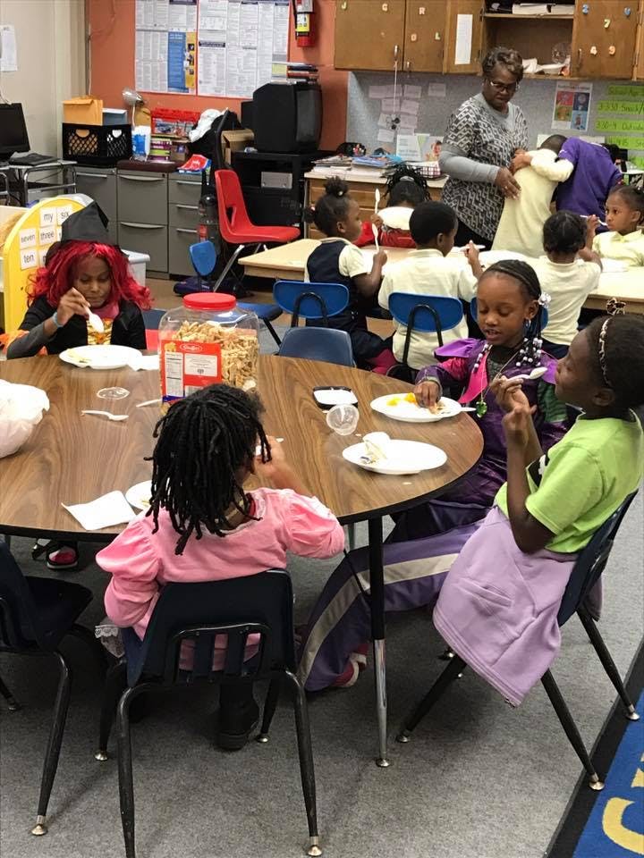 Children eating at tables in a classroom with an adult in the background