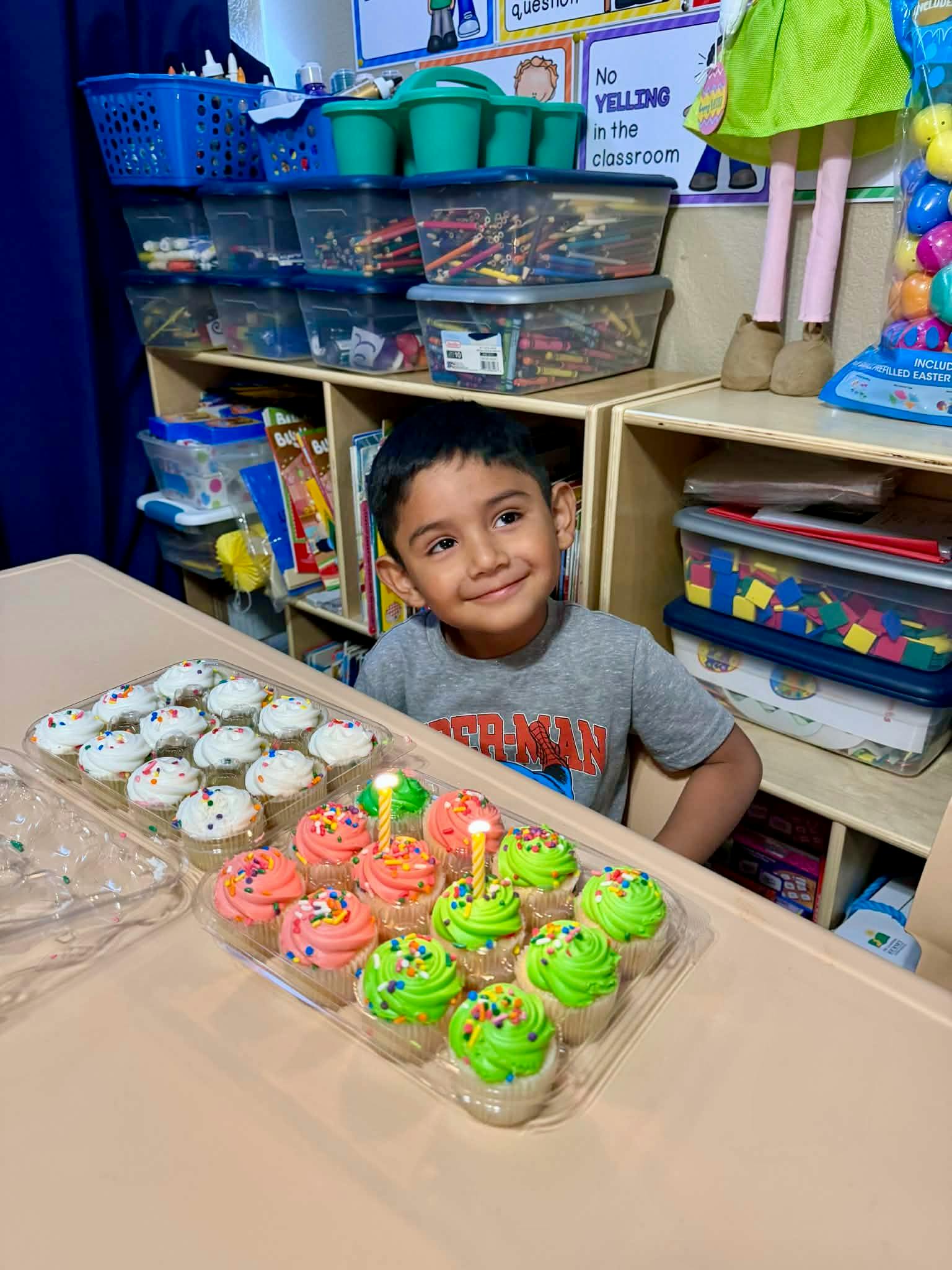 Child sitting at a table with two trays of cupcakes in a classroom